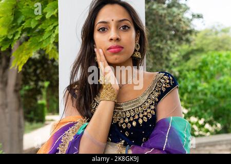 Jeune femme indienne de souche dans l'élégant traditionnel sari et bijoux debout regardant l'appareil photo dans le parc vert Banque D'Images