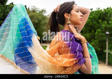 Vue latérale d'une jeune femme ethnique arrogante dans le sari traditionnel se tenant dans le parc parmi les arbres verts et tenant les cheveux tout en montrant de grandes boucles d'oreilles Banque D'Images