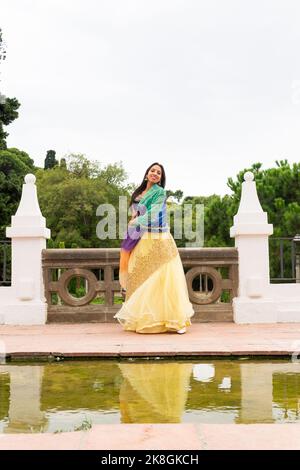 Corps complet de jeune femme indienne dans une élégante robe debout regardant la caméra dansant près de l'étang avec des arbres verts et des buissons en arrière-plan Banque D'Images