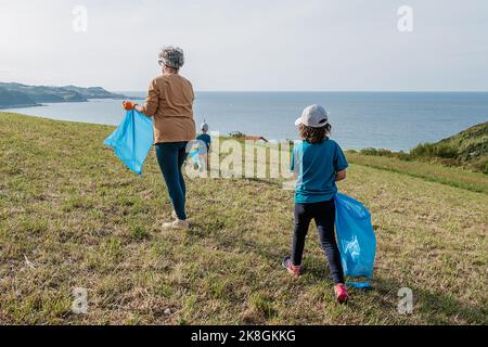 Des écoactivistes de différents âges dans les vêtements de sport avec des sacs de bac bleu collectant les déchets sur le vert herbeux colline près de mer bleu calme le jour ensoleillé Banque D'Images
