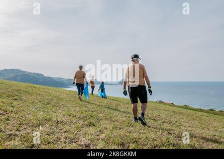 Des écoactivistes de différents âges dans les vêtements de sport avec des sacs de bac bleu collectant les déchets sur le vert herbeux colline près de mer bleu calme le jour ensoleillé Banque D'Images