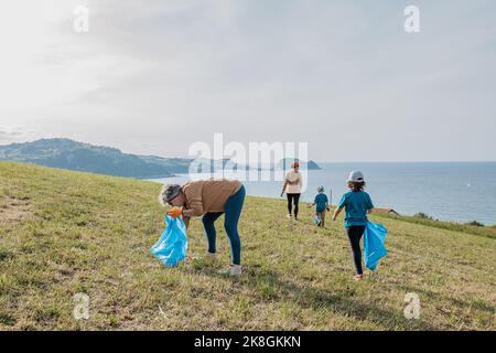 Des écoactivistes de différents âges dans les vêtements de sport avec des sacs de bac bleu collectant les déchets sur le vert herbeux colline près de mer bleu calme le jour ensoleillé Banque D'Images