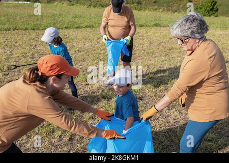 Famille écologique en casquettes cueillant les déchets à l'aide de pinces à thrash dans un sac poubelle bleu pour le recyclage sur une colline herbeuse près de la mer Banque D'Images