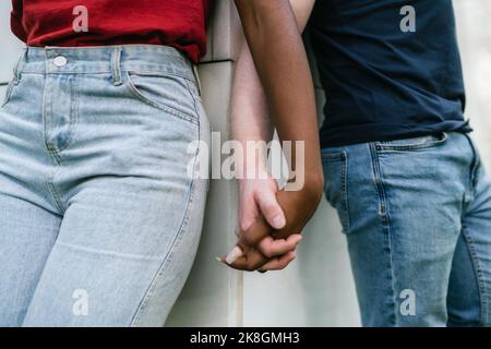 Cachez un petit ami et une petite amie multiracial méconnaissable en jeans et t-shirts inclinés sur un mur blanc et tenant les mains Banque D'Images