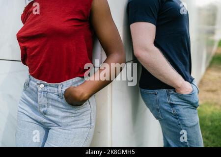 Cachez un petit ami et une petite amie multiracial méconnaissable en jeans et t-shirts inclinés sur un mur blanc avec les mains sur la poche Banque D'Images