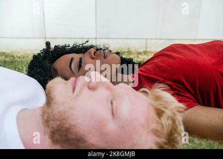 Vue d'en haut du côté d'un couple multiethnique non émotionnel allongé sur une pelouse verte dans le parc, près du mur de tuiles blanches tout en regardant vers le haut Banque D'Images