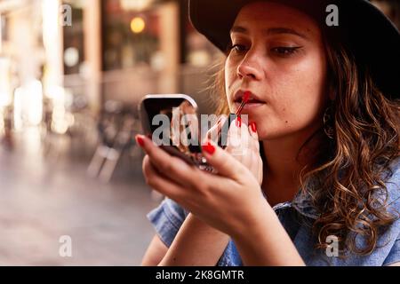 Jeune femme dans un chapeau regardant dans le miroir et se maillant rouge rouge rouge à lèvres tout en appliquant le maquillage sur la rue de Madrid, Espagne Banque D'Images