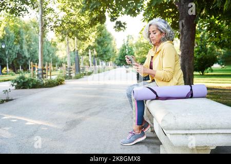 Vue latérale du corps de la femme sénior concentrée avec des cheveux courts gris dans les vêtements de sport assis sur un banc de pierre près du tapis de yoga roulé et de naviguer sur Internet v Banque D'Images