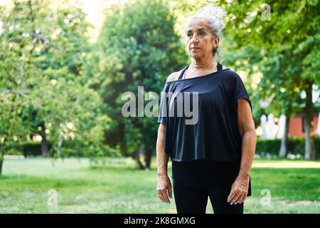 Femme âgée calme et peu émotive dans des vêtements de sport noirs avec une coiffure de pain sur une pelouse verte dans le parc au milieu des arbres foliaires et en regardant loin pendant que vous vous reposez Banque D'Images