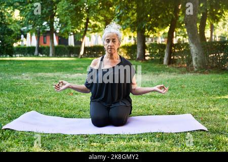Une femme sénieuse ferme les yeux et gante la mudra gyan tout en s'agenouillant sur un tapis et en méditant pendant la séance de yoga le jour d'été dans le parc Banque D'Images