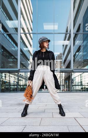 Jeune femme en vêtement d'extérieur élégant avec bonnet et lunettes se tenant à l'écart tout en tenant des fleurs orange contre un bâtiment moderne avec des murs en verre sur c Banque D'Images