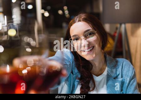Jeune femme gaie Manager dans des vêtements et des lunettes élégants et décontractés souriant et regardant l'appareil-photo tout en regardant des lunettes de vin avec un partenaire pendant les affaires Banque D'Images