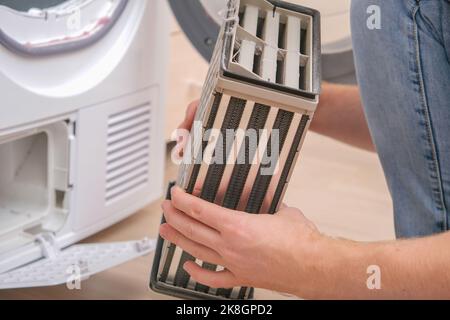 Machine à sécher avec un filtre plein de peluches, cheveux, poussière, laine après le cycle de séchage des serviettes, linge de lit. Machine à sécher blanche. Un homme sort un sécheur de radiateur sale. Banque D'Images