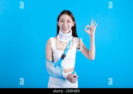 Belle jeune femme asiatique avec bras cassé dans une attelle douce souffrant d'un bras douloureux montrant le signe OK isolé sur fond bleu, concept d'assurance accident. Banque D'Images