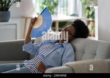 Femme Afro-américaine épuisée la femme au foyer se trouve sur le canapé stressé en raison de la température élevée Banque D'Images