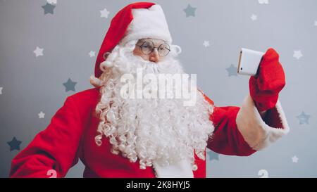 Portrait du Père Noël prenant le selfie avec l'appareil photo de smartphone sur fond gris pailleté, touchant l'écran et regardant l'appareil photo. Moern gadgets et concept de vacances. Banque D'Images