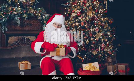 Le Père Noël est l'emballage de cadeaux dans le sac se préparer pour les célébrations du nouvel an près de l'arbre de Noël, les décorations et la cheminée sont visibles en arrière-plan. Banque D'Images