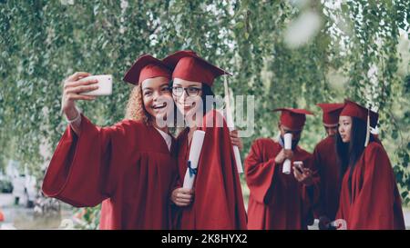 Les jolies femmes diplômées prennent le selfie avec le défilement du diplôme en utilisant le smartphone, les jeunes femmes posent avec d'autres étudiants dans les robes mobiles et parler en arrière-plan. Banque D'Images