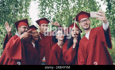 Des amis joyeux les étudiants diplômés prennent le selfie avec leur smartphone, les jeunes posent, montrent des gestes de main, font des visages amusants, sourient et rient. Banque D'Images