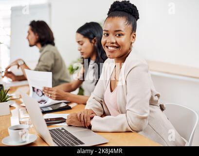 C'est ici que nous travaillons notre magie en équipe. Portrait d'une jeune femme d'affaires assise dans un bureau avec ses collègues en arrière-plan. Banque D'Images