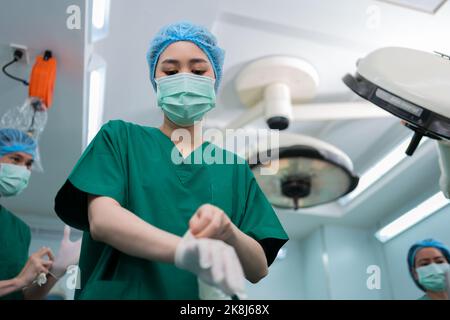 Portrait d'un chirurgien asiatique avec un masque médical debout et portant des gants médicaux dans une salle d'opération à l'hôpital. Équipe de chirurgiens professionnels. Hé Banque D'Images