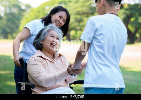 Une grand-mère asiatique heureuse utilise un fauteuil roulant avec sa fille et son petit-enfant dans le parc, le petit-fils est venu rendre visite à une grand-mère âgée et la main. Con Banque D'Images