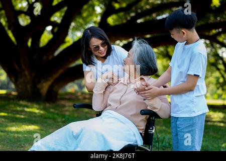 Une grand-mère asiatique heureuse utilise un fauteuil roulant avec sa fille et son petit-enfant dans le parc, le petit-fils est venu rendre visite à une grand-mère âgée et la main. Con Banque D'Images