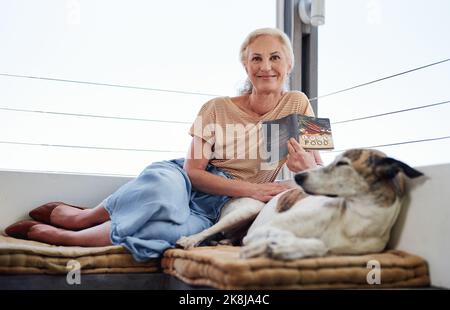 Il aime sa compagnie. Portrait en longueur d'une femme âgée attrayante lisant un livre tout en se relaxant sur le balcon avec son chien. Banque D'Images