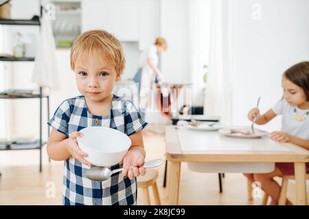 Portrait d'un petit garçon montrant son bol vide après le dîner. Sa sœur mange toujours en arrière-plan. Banque D'Images