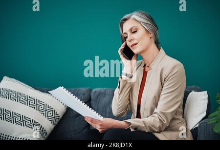 Je suis en train de lire les documents pendant que nous parlons. Une femme d'affaires attirante et mûre assise seule et utilisant son téléphone portable tout en lisant les documents dans elle Banque D'Images