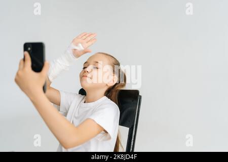 Positive blonde petite fille avec bras cassé enveloppé dans le bandage de plâtre blanc parler appel vidéo sur le téléphone portable assis dans la chaise sur blanc isolé Banque D'Images