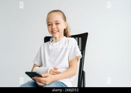 Portrait de la petite fille blonde positive avec le bras cassé enveloppé dans le bandage de plâtre blanc souriant regardant l'appareil photo tenant le téléphone mobile assis Banque D'Images