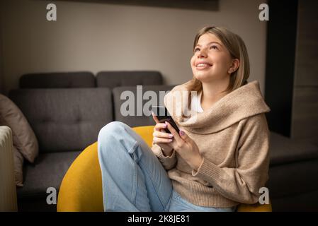 Bonne femme regardant des vidéos ou appréciant du contenu de divertissement sur un mobile assis sur le canapé dans la maison. Banque D'Images