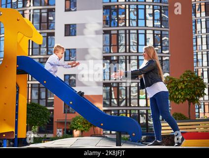 Jolie maman qui attrape un enfant, qui se glisse dans la cour moderne des immeubles résidentiels en hauteur de la ville. Mère attrapant un petit fils en se tenant près du toboggan sur l'aire de jeux. Concept de la parentalité. Banque D'Images