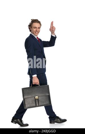 homme d'affaires excité avec une valise pointant les doigts dans l'air et saluant tout en marchant isolé devant un fond blanc dans le studio Banque D'Images