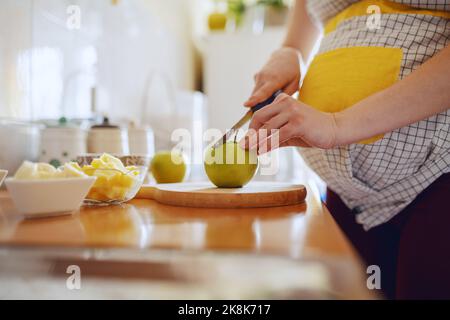 Gros plan de la femme enceinte de race blanche en tablier coupant la pomme verte dans la cuisine. Banque D'Images