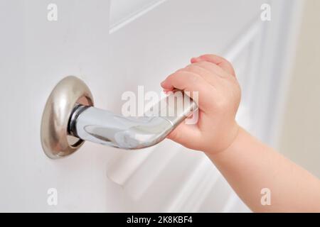 Tout-petit ouvre la porte maintenant la poignée de porte, la main de l'enfant en gros plan. Porte en bois blanc, poignée de porte en métal et main de bébé Banque D'Images