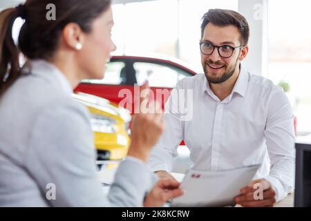 Sourire le client assis à la table avec le vendeur de voiture, tenant l'offre et écoutant le vendeur de voiture au sujet des commodités. Banque D'Images