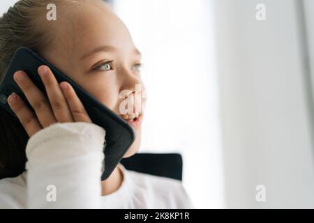 Gros plan d'une petite fille blonde gaie avec un bras cassé enveloppé dans un bandage en plâtre parlant smartphone, souriant regardant loin sur fond blanc. Banque D'Images