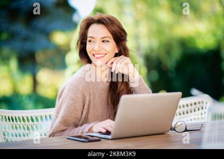 Femme d'âge moyen confiante avec ordinateur portable sur le balcon. Jolie femme vêtue d'un pull et souriante. Banque D'Images