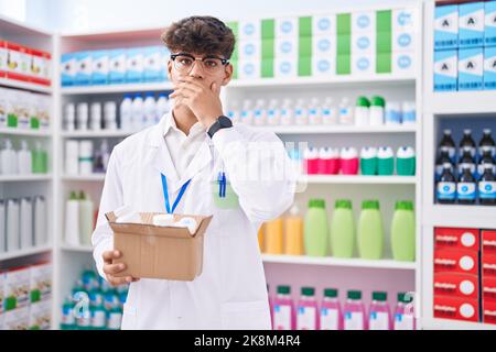 Adolescent hispanique travaillant à pharmacie drugstore tenant boîte avec des pilules couvrant la bouche avec la main, choqué et peur pour l'erreur. Expression surprise Banque D'Images