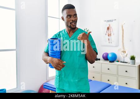 Un jeune kinésithérapeute africain tenant un rouleau en mousse à la clinique pointant le pouce vers le côté souriant heureux avec la bouche ouverte Banque D'Images