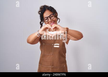 Femme d'âge moyen portant un tablier sur fond blanc souriant dans l'amour faisant le symbole de coeur forme avec les mains. Concept romantique. Banque D'Images