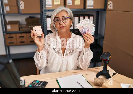 Femme d'âge moyen avec cheveux gris travaillant à la petite entreprise ecommerce tenant la banque de porc et zloty détendu avec l'expression sérieuse sur le visage. Simple et n Banque D'Images