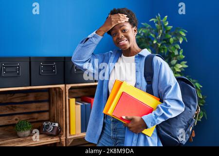 Femme afro-américaine portant un sac à dos étudiant et tenant des livres stressés et frustrés avec la main sur la tête, surpris et fâché visage Banque D'Images
