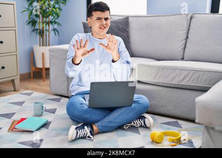 Personne non binaire étudiant en utilisant ordinateur portable assis sur le sol peur et terrifié avec l'expression de peur arrêter le geste avec les mains, criant dans les s. Banque D'Images
