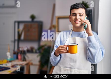 Jeune artiste homme non binaire parlant sur le smartphone boire du café au studio d'art Banque D'Images