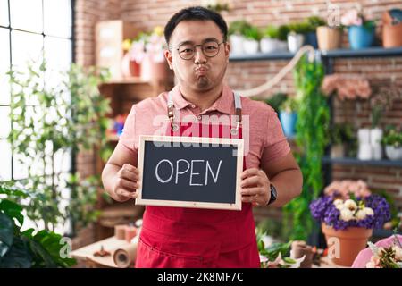 Jeune homme chinois travaillant chez le fleuriste tenant des joues ouvertes avec le visage drôle. Bouche gonflée avec de l'air, attrapant l'air. Banque D'Images