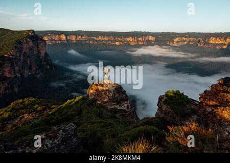 Femme randonneur se tient sur une pagode rocheuse ihigh dans les Blue Mountains au-dessus d'une vallée de brouillard brumeux rempli comme les premiers rayons du soleil frappent les falaises d'escarpement et Banque D'Images