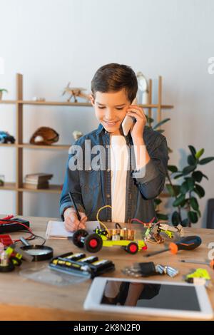 garçon souriant parlant sur smartphone et écrivant dans ordinateur portable à proximité de pièces mécaniques et tablette numérique floue, image de stock Banque D'Images